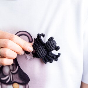 May include: A black crocheted spider with eight legs. The spider is being held by a hand in front of a white shirt with a Halloween-themed design.