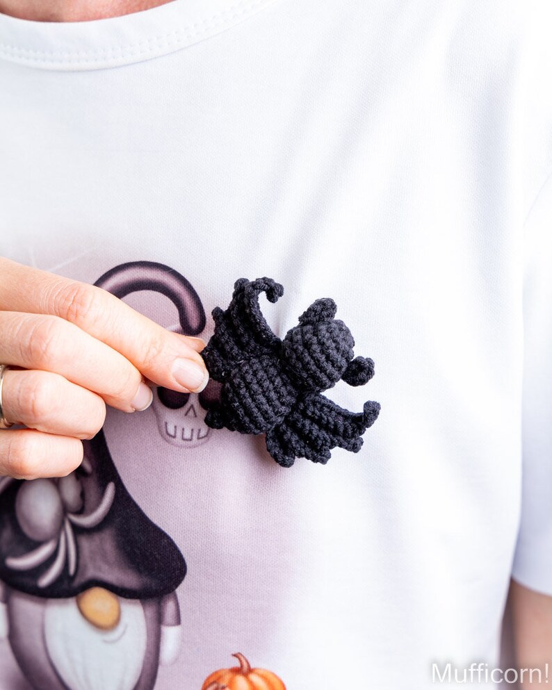 May include: A black crocheted spider with eight legs. The spider is being held by a hand in front of a white shirt with a Halloween-themed design.