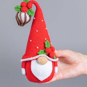 May include: A red crocheted gnome with a white beard and a red hat decorated with green leaves and red berries. The hat has white pearl beads around the brim.