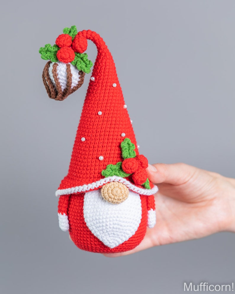 May include: A red crocheted gnome with a white beard and a red hat decorated with green leaves and red berries. The hat has white pearl beads around the brim.