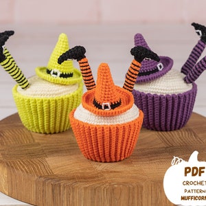May include: Three crocheted witch cupcake toppers with legs sticking up in the air. The cupcakes are yellow, orange, and purple. The toppers are wearing black witch hats. The cupcakes are sitting on a wooden surface. The text "PDF CROCHET PATTERN MUFFICORN" is visible in the lower right corner of the image.