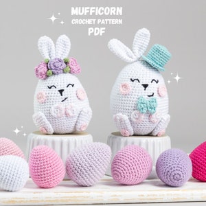 Könnte beinhalten: Zwei gehäkelte Osterhasen-Plüschtiere, eines mit einer Blumenkrone und das andere mit einem blauen Hut und einer Fliege. Die Hasen sitzen auf einer weißen Oberfläche mit sechs gehäkelten Ostereiern in Rosa, Lila und Weiß.