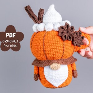 Puede incluir: Un gnomo con forma de calabaza tejido a ganchillo, con cuerpo naranja, barba blanca y bufanda marr&oacute;n. El gnomo tiene un sombrero blanco y sostiene un asa marr&oacute;n. La imagen incluye el texto "PDF CROCHET PATTERN".