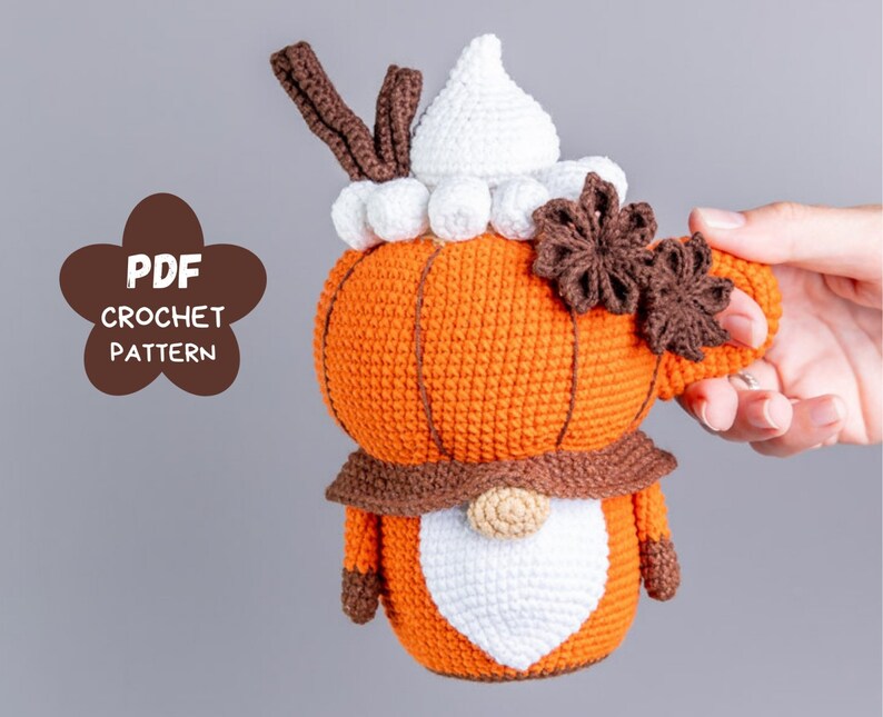 Puede incluir: Un gnomo con forma de calabaza tejido a ganchillo, con cuerpo naranja, barba blanca y bufanda marr&oacute;n. El gnomo tiene un sombrero blanco y sostiene un asa marr&oacute;n. La imagen incluye el texto "PDF CROCHET PATTERN".