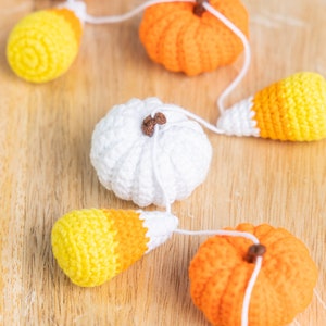 May include: A crocheted garland featuring alternating orange pumpkins, white pumpkins, and yellow and orange candy corn. The garland is strung together with white yarn.