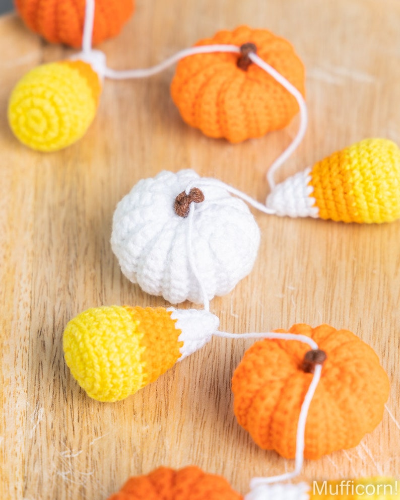 May include: A crocheted garland featuring alternating orange pumpkins, white pumpkins, and yellow and orange candy corn. The garland is strung together with white yarn.