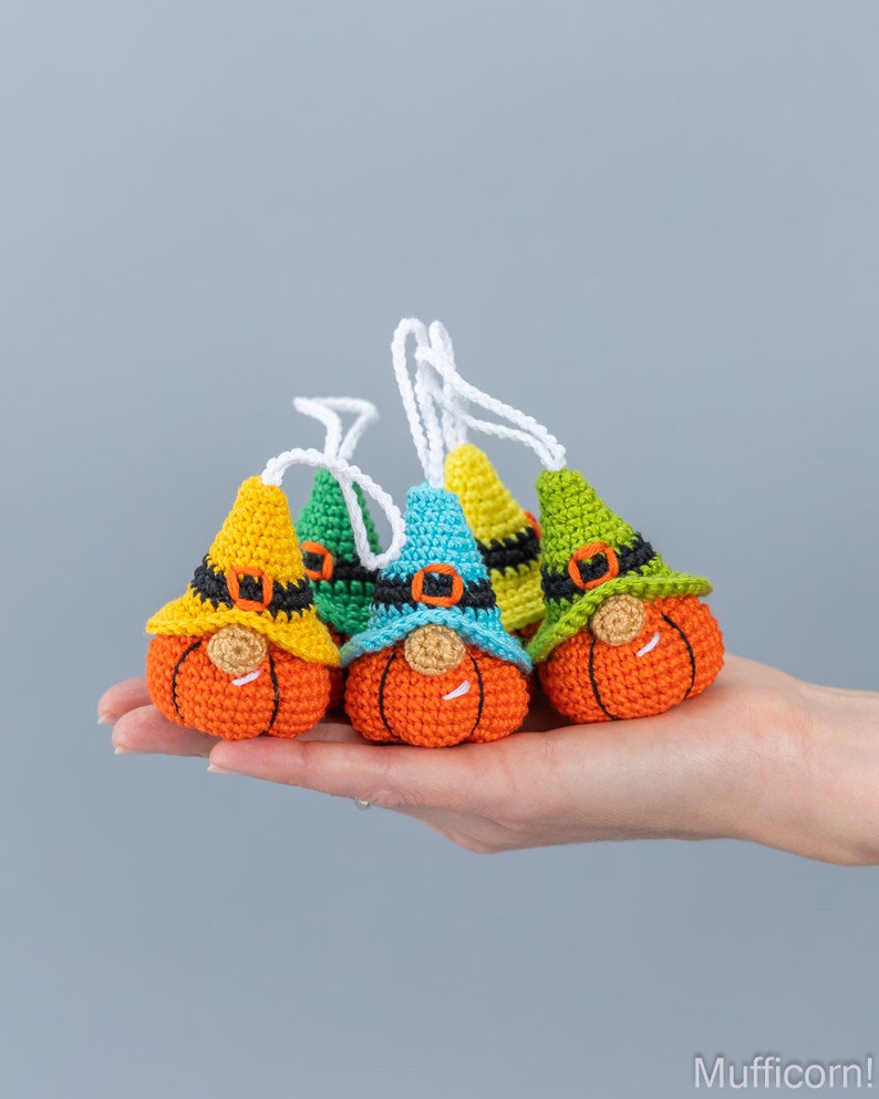 May include: Five crocheted pumpkin gnomes with different colored hats. The gnomes are orange with black eyes and a black belt. The hats are yellow, green, blue, and green.