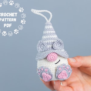 May include: Crochet pattern PDF for a gray and pink gnome ornament with a striped hat and paw print details.