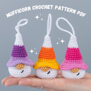 May include: Three crocheted gnome ornaments with different colored hats. The gnomes are white with black eyes and a red nose. The hats are purple, orange and yellow, and pink. The gnomes are hanging from a hand.