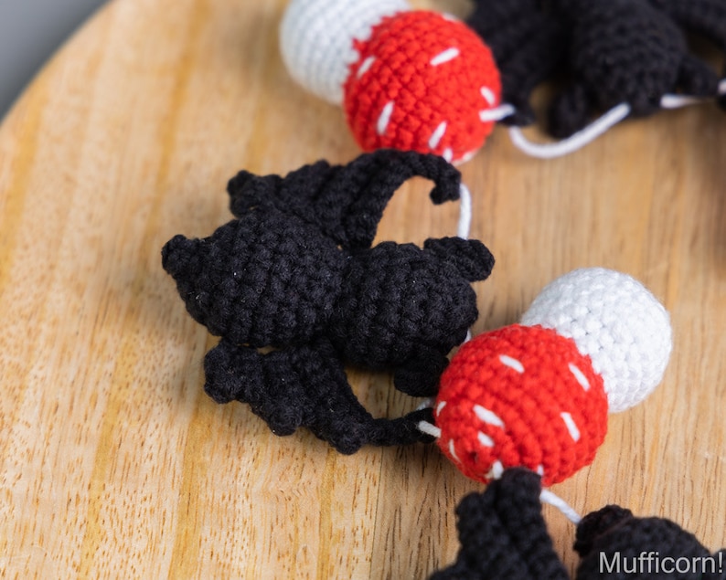 May include: A crocheted garland with alternating black bats and red and white mushrooms. The garland is strung together with white yarn.