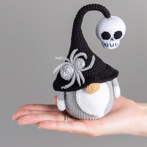 May include: A crocheted gnome with a black hat, a white skull, and a gray spider on the hat. The gnome is wearing a gray and white beard.