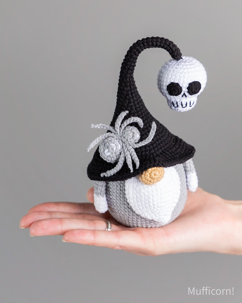 May include: A crocheted gnome with a black hat, a white skull, and a gray spider on the hat. The gnome is wearing a gray and white beard.