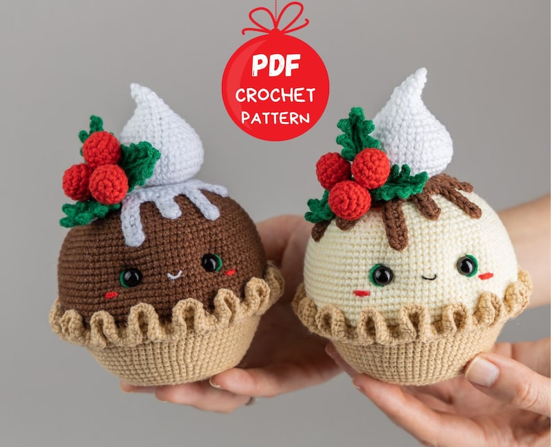 May include: Two crocheted Christmas pudding ornaments with smiling faces. The ornaments are decorated with green holly leaves and red berries. One ornament is brown and the other is white.