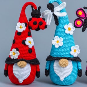 May include: Three crocheted gnomes with different colored hats and decorations. The gnome on the left has a red hat with a ladybug on top and white flowers. The gnome in the middle has a blue hat with a dragonfly on top and white flowers. The gnome on the right has a pink hat with a butterfly on top.