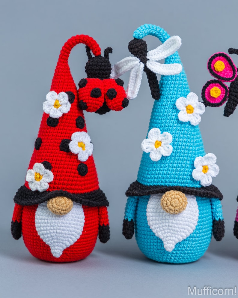 May include: Three crocheted gnomes with different colored hats and decorations. The gnome on the left has a red hat with a ladybug on top and white flowers. The gnome in the middle has a blue hat with a dragonfly on top and white flowers. The gnome on the right has a pink hat with a butterfly on top.