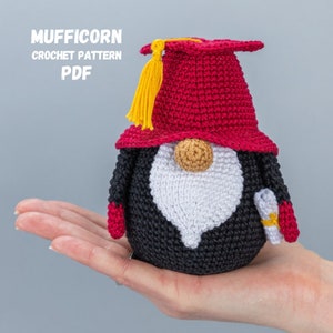 Puede incluir: Un gnomo de crochet que lleva una gorra de graduación roja con un borla amarilla y una túnica de graduación negra y blanca. El gnomo sostiene un rollo en su mano.