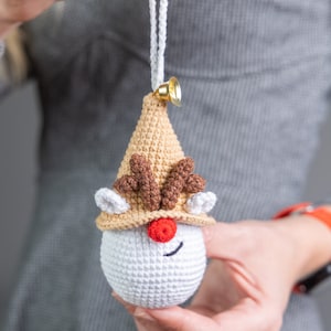 May include: A crocheted reindeer ornament with a white body, brown antlers, and a tan hat. The ornament has a red nose and black eyes. It is hanging from a white string with a gold bell.