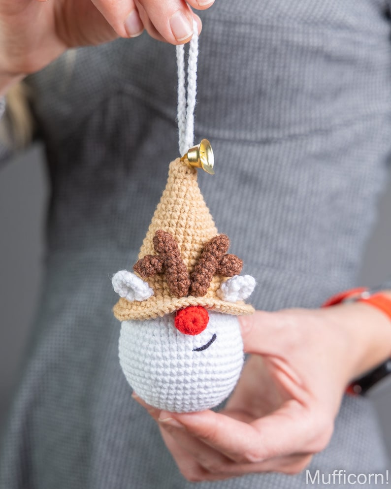 May include: A crocheted reindeer ornament with a white body, brown antlers, and a tan hat. The ornament has a red nose and black eyes. It is hanging from a white string with a gold bell.