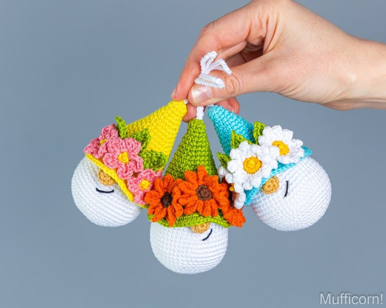 May include: Three crocheted gnome ornaments with white faces, black eyes, and red noses. Each gnome has a different colored hat with flowers: pink and yellow flowers, orange flowers, and white flowers.
