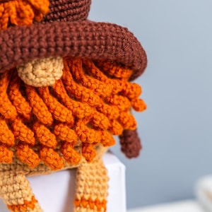 May include: Close-up of a crocheted gnome with a brown hat, orange beard, and tan legs. The gnome is sitting on a white surface.