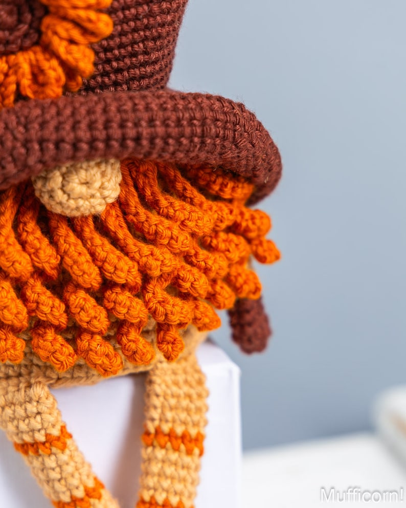 May include: Close-up of a crocheted gnome with a brown hat, orange beard, and tan legs. The gnome is sitting on a white surface.