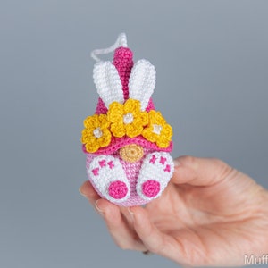 May include: A pink and white crocheted Easter gnome with a yellow flower crown and bunny ears.