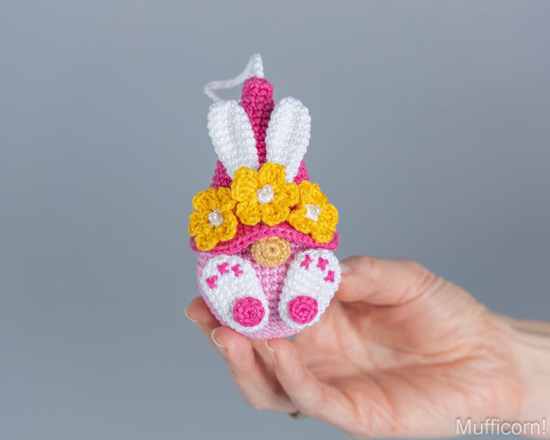 May include: A pink and white crocheted Easter gnome with a yellow flower crown and bunny ears.