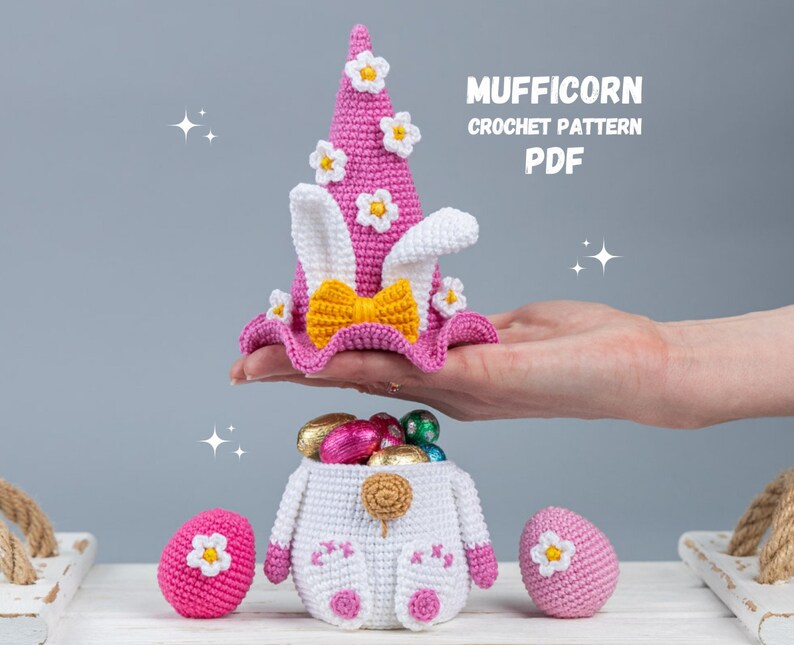 May include: A pink crocheted unicorn hat with white ears and yellow bow, a white crocheted basket filled with chocolate eggs, and two pink crocheted eggs. The text "MUFFICORN CROCHET PATTERN PDF" is displayed above the hat.