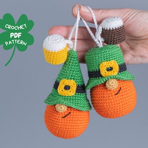 Op de afbeelding: Twee gehaakte kabouter ornamenten met groene hoeden, oranje gezichten en zwarte riemen. Elk ornament heeft een kleine gehaakte bierbeker.