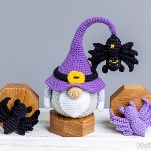 Puede incluir: Un gnomo de crochet morado con un sombrero de bruja con un borde negro y una hebilla amarilla. El gnomo sostiene un murci&eacute;lago de crochet negro. Hay dos murci&eacute;lagos de crochet m&aacute;s, uno negro y uno morado, sobre soportes de madera.