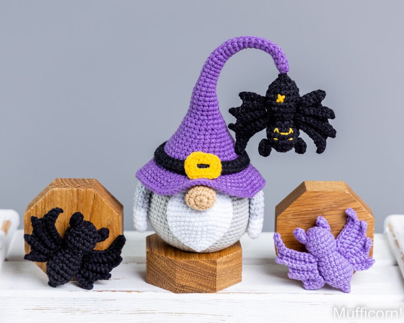 Puede incluir: Un gnomo de crochet morado con un sombrero de bruja con un borde negro y una hebilla amarilla. El gnomo sostiene un murci&eacute;lago de crochet negro. Hay dos murci&eacute;lagos de crochet m&aacute;s, uno negro y uno morado, sobre soportes de madera.