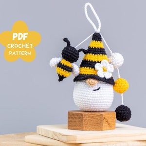 Puede incluir: Patrón de crochet PDF para un adorno de gnomo abeja. El gnomo es blanco con un sombrero a rayas negro y amarillo, una flor blanca y una abeja negra y amarilla. El gnomo está sentado sobre una base de madera.