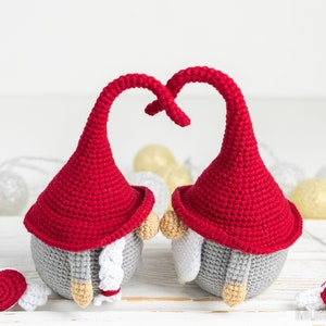 May include: Two crocheted gnomes with red hats and gray bodies are facing each other with their hats forming a heart shape. They are holding small red hearts with white wings.