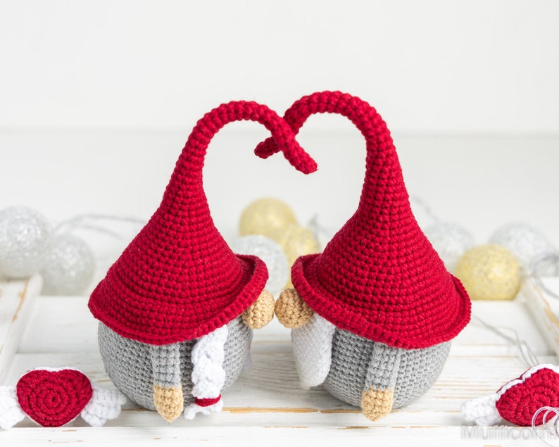 May include: Two crocheted gnomes with red hats and gray bodies are facing each other with their hats forming a heart shape. They are holding small red hearts with white wings.