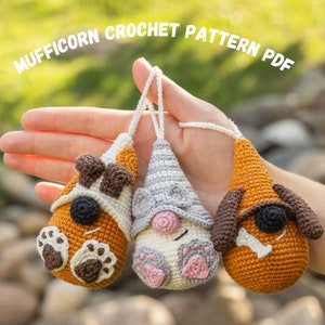 May include: Three crocheted amigurumi toys in the shape of dogs. The toys are brown, white, and gray with black eyes and noses. The toys are being held in a person's hand. The toys are part of a crochet pattern PDF.