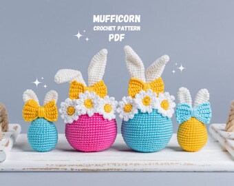 May include: Four crocheted bunny-shaped Easter eggs in blue, pink, yellow, and white with daisy accents. The eggs are arranged on a white wooden surface. The text "MUFFICORN CROCHET PATTERN PDF" is displayed above the eggs.