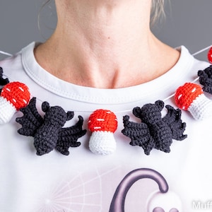 May include: A white shirt with a crocheted garland of black bats and red and white mushrooms. The garland is worn as a necklace.