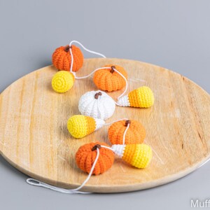 May include: A garland of crocheted pumpkins and candy corn on a wooden surface. The garland is made of white yarn and features orange, white, and yellow pumpkins and candy corn.