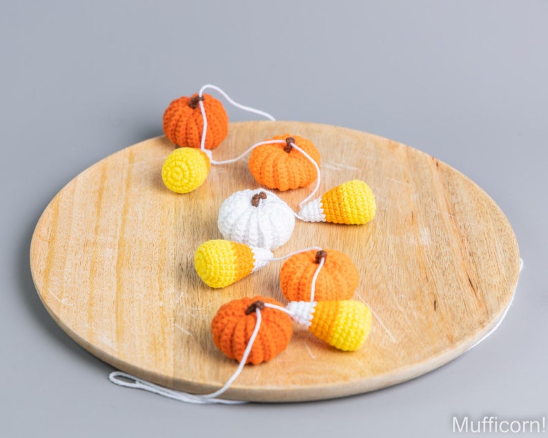 May include: A garland of crocheted pumpkins and candy corn on a wooden surface. The garland is made of white yarn and features orange, white, and yellow pumpkins and candy corn.