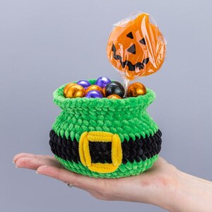 May include: A small, green, crocheted cauldron-shaped basket featuring a black band with a yellow buckle. The basket is filled with small, round, foil-wrapped candies in orange, purple, and black, and a single orange pumpkin-shaped lollipop.