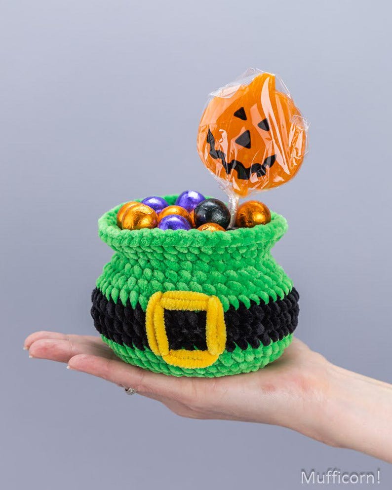 May include: A small, green, crocheted cauldron-shaped basket featuring a black band with a yellow buckle. The basket is filled with small, round, foil-wrapped candies in orange, purple, and black, and a single orange pumpkin-shaped lollipop.