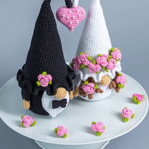 May include: Two crocheted gnomes dressed as a bride and groom. The bride gnome is wearing a white dress with pink flowers and a veil. The groom gnome is wearing a black suit with a white bow tie. They are holding a pink heart.