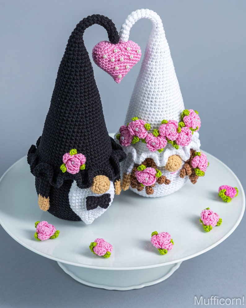 May include: Two crocheted gnomes dressed as a bride and groom. The bride gnome is wearing a white dress with pink flowers and a veil. The groom gnome is wearing a black suit with a white bow tie. They are holding a pink heart.