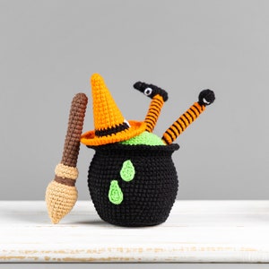 May include: A crocheted witch doll with orange and black striped legs, a black cauldron, and a green pointed hat. The doll is sitting in the cauldron with its legs sticking out. The doll is holding a brown and orange crocheted broom.