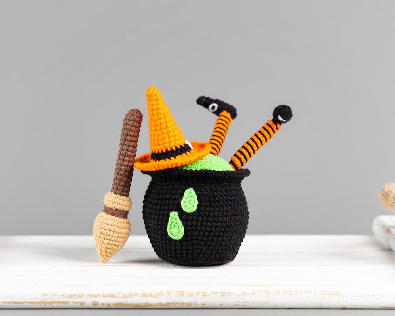 May include: A crocheted witch doll with orange and black striped legs, a black cauldron, and a green pointed hat. The doll is sitting in the cauldron with its legs sticking out. The doll is holding a brown and orange crocheted broom.