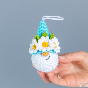 May include: A crocheted gnome ornament with a blue hat, white flowers, and green leaves. The gnome has a white face with black eyes and a brown nose.