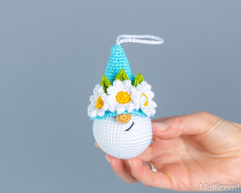 May include: A crocheted gnome ornament with a blue hat, white flowers, and green leaves. The gnome has a white face with black eyes and a brown nose.