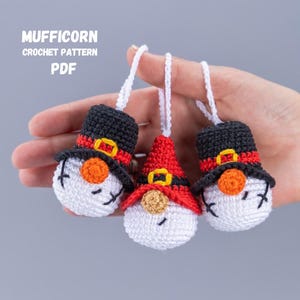 Könnte beinhalten: Drei gehäkelte Schneemann-Anhänger mit schwarzen Hüten, roten und weißen Gesichtern und gelben Akzenten. Die Anhänger hängen an weißen Schnüren und werden in einer Hand gehalten.