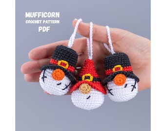 May include: Three crocheted snowman ornaments with black hats, red and white faces, and yellow accents. The ornaments are hanging from white strings and are held in a hand.