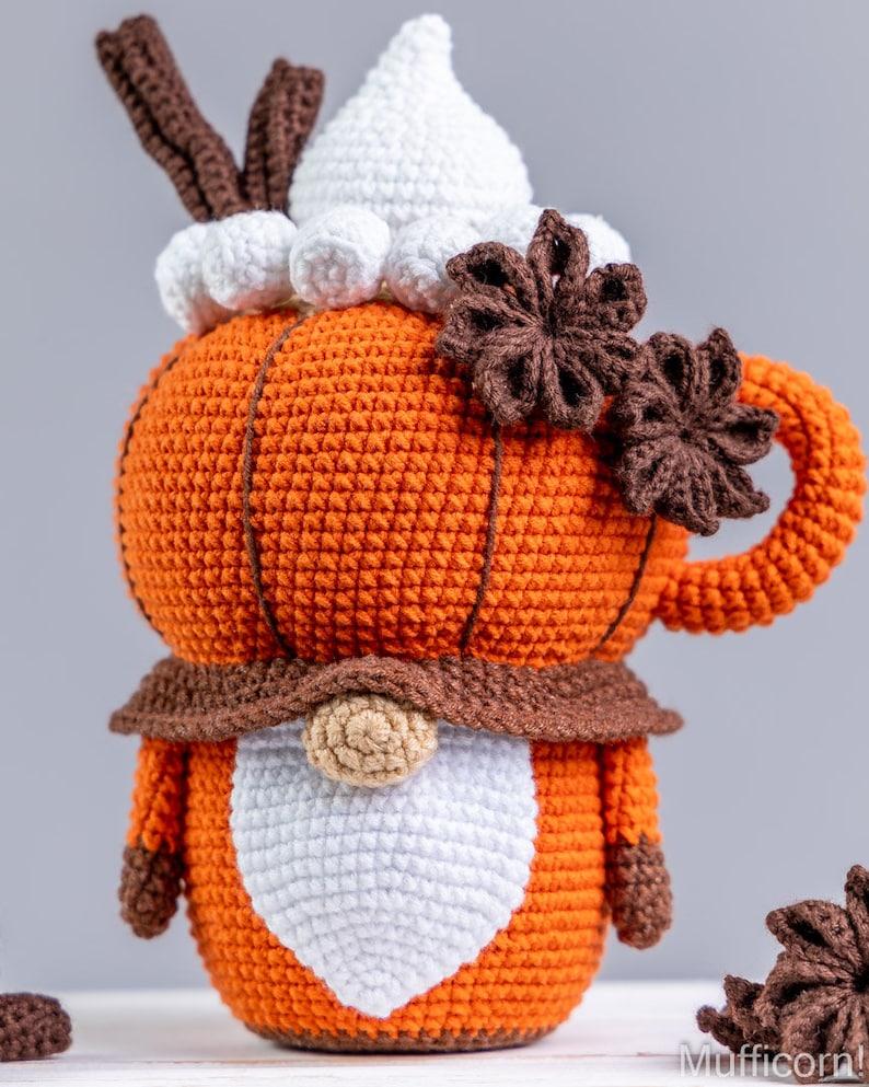 Puede incluir: Un gnomo con forma de calabaza naranja tejido a ganchillo, con barba y sombrero blancos, detalles marrones y un asa de taza. La calabaza est&aacute; adornada con an&iacute;s estrellado marr&oacute;n y una cobertura de crema batida blanca. Un art&iacute;culo decorativo con tem&aacute;tica oto&ntilde;al.